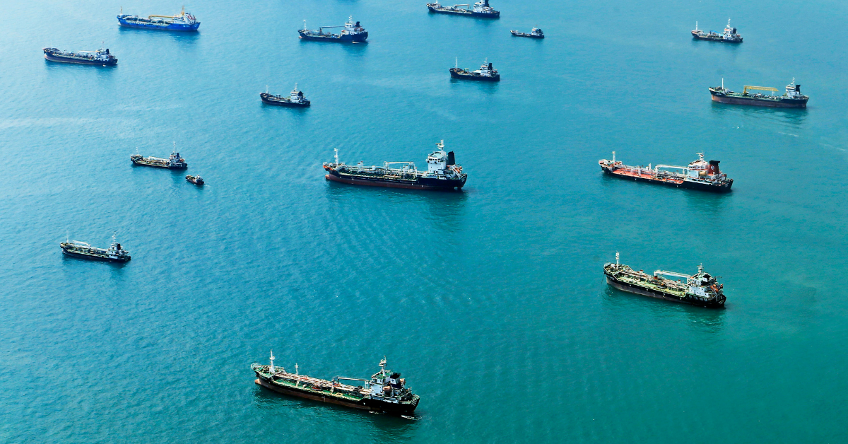 A fleet of large cargo ships are anchored in a vast expanse of blue water, viewed from above.